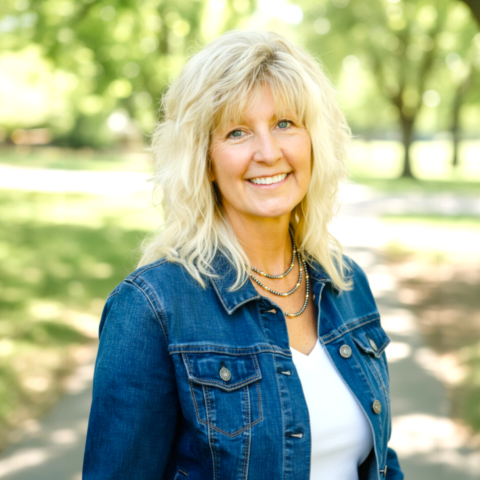 Woman with blonde hair wearing a blue denim jacket and silver and gold necklaces in a park setting