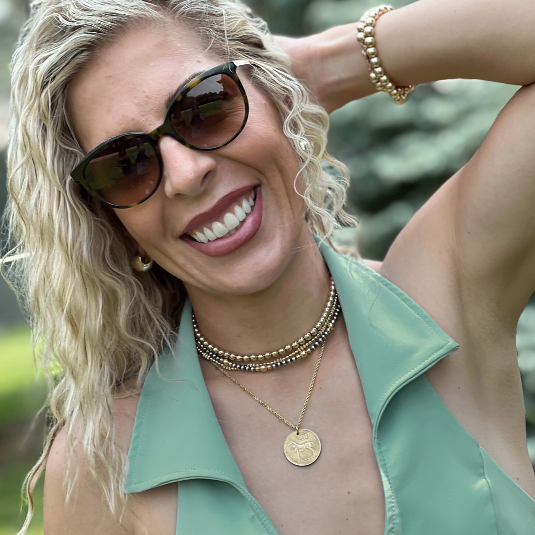 Woman wearing sunglasses and a green top and gold jewelry with a blurred natural background