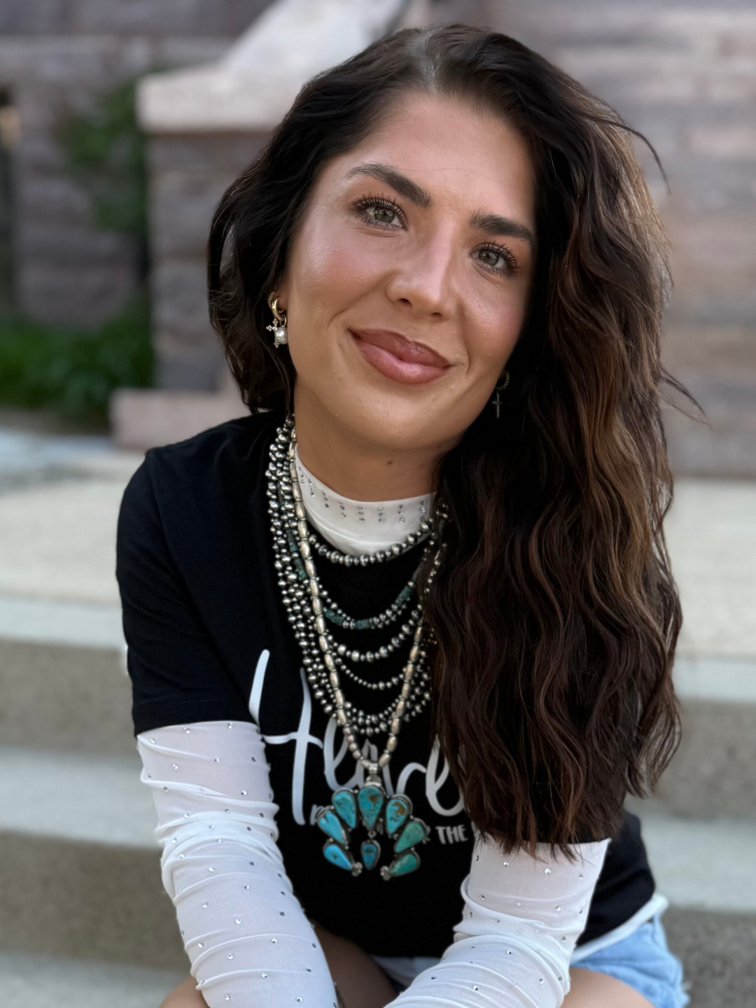 Woman sitting on steps wearing a black shirt and multiple silver necklaces.