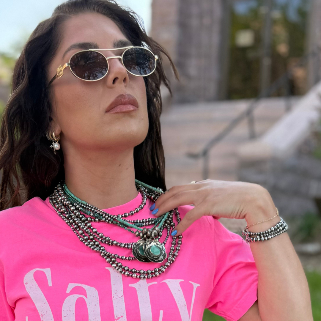 Woman wearing sunglasses and a pink shirt with visible text and silver jewelry, outdoors.